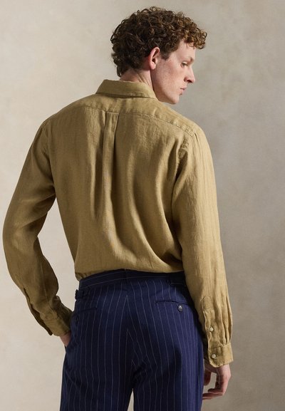 Lniana koszula w oliwkowej zieleni z tylnym karczkiem i rękawami zakończonymi mankietami na guziki. Połączona z ciemnoniebieskimi spodniami w cienkie prążki, prezentującymi dopasowany krój.