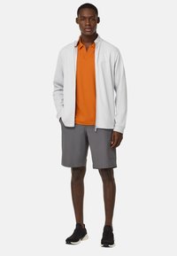 Chaqueta de cremallera gris claro sobre una polo naranja, combinada con pantalones cortos grises y zapatillas deportivas negras. Tejido suave con un ajuste casual.