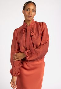 Rote strukturierte Bluse mit gebundenem Kragen und elastischen Manschetten, kombiniert mit einem passenden einfarbigen Rock. Verfügt über einen dezenten Glanz und voluminöse Ärmel.