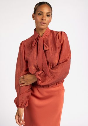 Rode blouse van textuur met een geknoopte hals en elastische manchetten, gecombineerd met een bijpassende effen rok. Heeft een subtiele glans en volumineuze mouwen.