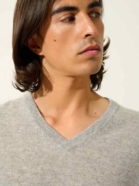 Ljusgrå v-ringad tröja gjord av mjukt tyg, med en ren halsringning och subtila stickdetaljer. Hudton varm, hår mörkt och vågigt.