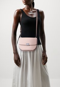 Růžová kožená crossbody taška s kulatým tvarem, stříbrným kováním a klopovým uzavíráním. Modelka má na sobě černý top na ramínka a bílou sukni.