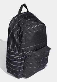 Svart Adidas ryggsäck med vitt upprepat logomönster, framficka med dragkedja och handtag för bärande på toppen, mot en ljus bakgrund.