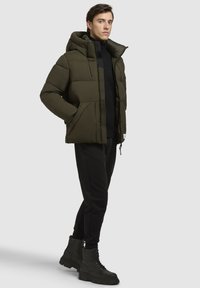 Chaqueta de plumas verde oliva oscuro con capucha, que presenta secciones acolchadas, cremallera frontal y bolsillos laterales, combinada con joggers negros y botas de combate.