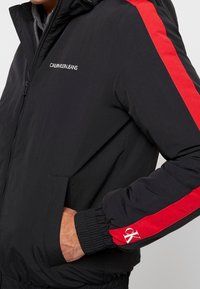 Svart jacka med röda sidostrippar, försedd med dragkedja, elastiska muddar och "Calvin Klein Jeans"-logotyp på bröstet.