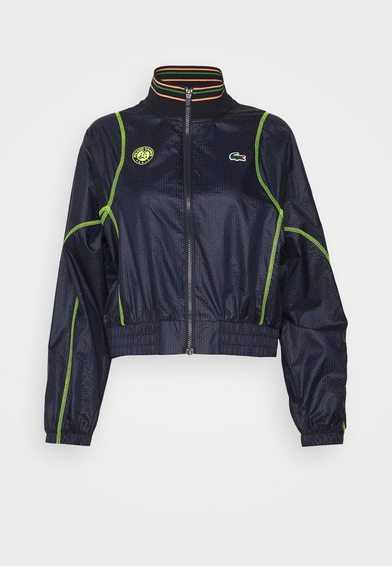 Veste courte marine avec fermeture éclair à l'avant, surpiqûres vertes et col rayé. Présente un patch logo sur la poitrine et des poignets élastiques.