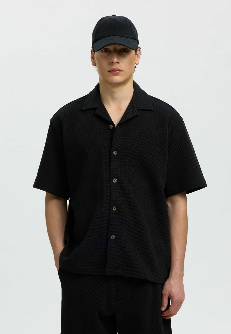 Jeune homme portant une chemise noire à manches courtes boutonnée, une casquette noire et un pantalon noir assorti sur un fond clair uni.
