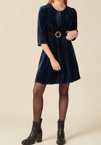 Vestido de terciopelo azul marino con mangas tres cuartos y un escote en forma de ojal, ceñido a la cintura con un cinturón negro que presenta una hebilla redonda.