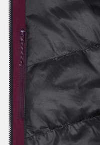 Bordeauxfärgad isolerad jacka med insidan i ett slätt, svart tyg med ett diagonalt quiltat mönster och en dragkedja på vänster sida.