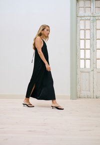 Femme marchant à l'intérieur portant une robe longue noire sans manches avec une fente latérale et des sandales noires à petits talons kitten près d'une porte blanche vintage.