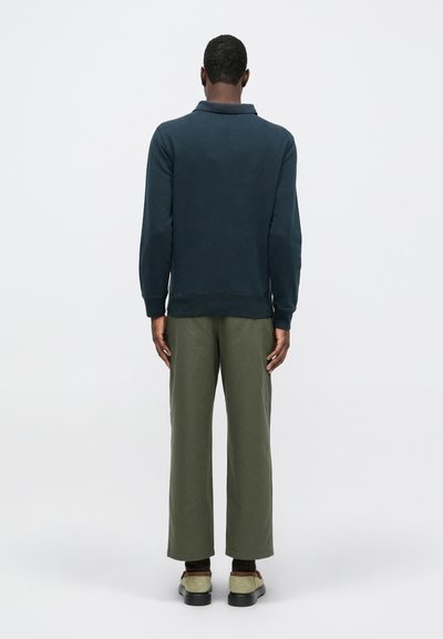 Pulover bleu marin cu guler, mâneci lungi și manșete cu volum, asortat cu pantaloni lejeri de culoare verde măslin și încălțăminte în contrast.