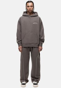 Felpa grigia con tasca a marsupio, abbinata a pantaloni tuta grigi coordinati con cuciture bianche. Il materiale sembra morbido e informale.