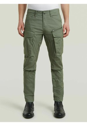 Uomo che indossa pantaloni cargo verde oliva con più tasche e stivali neri, in piedi davanti a uno sfondo chiaro e semplice.