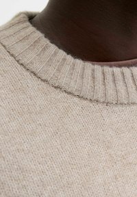 Beige stickad tröja med ribbad halsringning och mjuk textur. Närbild av tygdetaljen visar fina stygn och naturliga fibrer.