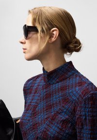 Rollkragenoberteil aus strukturiertem dunkelrot und blauem Karomuster mit hohem Kragen. Große schwarze Sonnenbrille. Haare im tiefen Dutt gestylt. Schwarze Handtasche sichtbar.