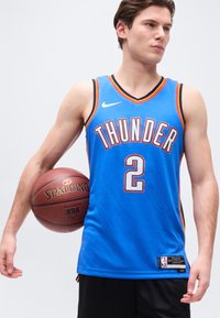 Jeune homme tenant un ballon de basketball sous le bras, portant un maillot bleu des Thunder avec le numéro 2, regardant sur le côté sur un fond uni.