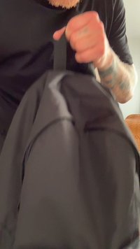 Mochila preta feita de um tecido durável, com uma superfície suave e alças ajustáveis. Uma mão está a segurar a pega superior.