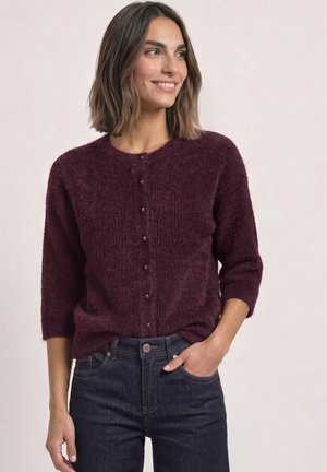 Femme souriante portant un pull violet foncé à boutons, duveteux, avec des manches trois-quarts et un jean bleu foncé, main dans la poche, regardant sur le côté.