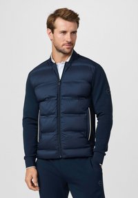 Chaqueta acolchada azul marino con cierre de cremallera frontal, diseño acolchado en el torso, mangas lisas y detalles laterales de color claro. Puños acanalados.