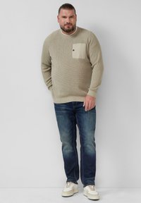 Beżowy sweter o teksturze, z kieszenią na piersi, w połączeniu z niebieskimi dżinsami i białymi trampkami. Sweter ma okrągły dekolt i ściągacze przy mankietach.