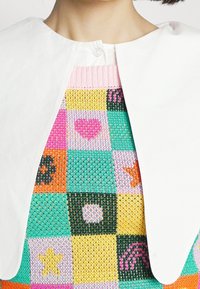 Haut en tricot avec un motif à carreaux multicolore featuring des cœurs, des étoiles et des fleurs ; col blanc avec fermeture à bouton.