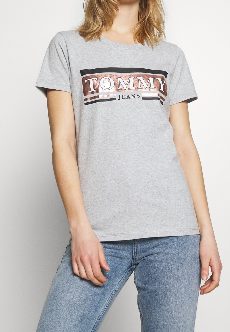 Osoba oblečená v světle šedém tričku Tommy Jeans s růžově zlatým a černým logem, doplněná modrými džíny, stojící na bílém pozadí.