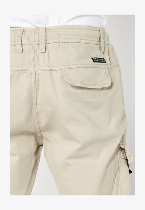 Beige cargo pants with an elastic waistband, a back pocket, and stitching details. Material appears durable and smooth in texture.