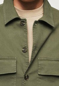 Oliivinvihreä napillinen takki, jossa on kaulus, kaksi rintataskua ja ruskeat napit. Kangas näyttää sileältä ja siinä on hienovarainen tekstuuri.