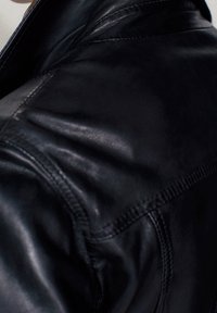 Gros plan de l'épaule d'une veste en cuir noire montrant des coutures détaillées et un matériau à texture lisse.