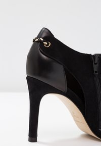 Chaussure à talon haut noire en combinaison de cuir lisse et de suède. Présente une fermeture éclair sur le côté et un détail tressé avec des accents dorés sur le talon.