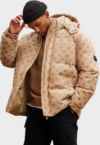 Beige prošívaná bunda s kapucí s hexagonálním vzorem a zlatými detaily. Vnitřní strana je podšitá hnědým materiálem, nošena přes hnědou mikinu.