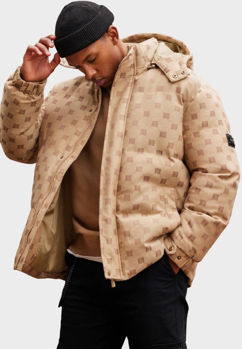 Beige prošívaná bunda s kapucí s hexagonálním vzorem a zlatými detaily. Vnitřní strana je podšitá hnědým materiálem, nošena přes hnědou mikinu.