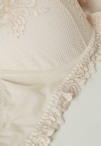 Sujeta beige claro con detalles de encaje texturizado y volantes. La tela presenta un patrón en cuadrícula, complementado con acentos de bordado floral.