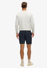 Sudadera gris claro de manga larga, combinada con pantalones cortos de carga azul marino que tienen bolsillos laterales. Zapatillas beige y calcetines de color claro completan el conjunto.
