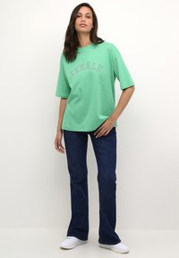 Mintgroene katoenen t-shirt met "EXHALE" in contrasterende letters, gecombineerd met donkerblauwe flared jeans en witte sneakers.