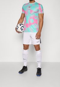 Nike-färgglad sporttröja i turkos och rosa, matchad med vita shorts och svarta fotbollsskor. Håller en mönstrad fotboll.