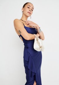 Navy blue satin dress with spaghetti straps, ruffled side detail, and thigh slit, paired with a cream-colored pouch bag.
