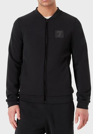Sweat à zip noir avec un col rond, des poches latérales et un tissu texturé. Il présente un petit écusson avec logo sur la poitrine.