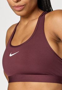 Bordeaux sportbh van glad materiaal met een brede band en racerback ontwerp; heeft een wit Nike-logo op de voorkant.