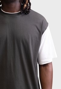 Tricou scurt de culoare gri închis, purtat peste un tricou alb cu mânecă lungă, prezentând o croială lejeră și un material neted, texturat.