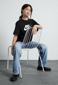 Fiatal személy hátradőlve ül egy fehér fém széken, fekete Nike pólót, világoskék farmernadrágot és fekete csizmát visel.