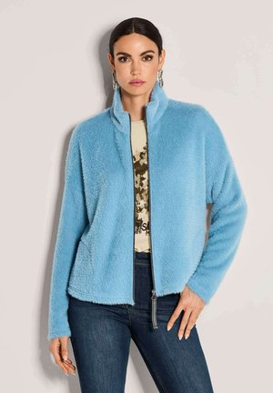 Vrouw in een blauwe fuzzy zip-up jas, met een beige T-shirt met print, donkere spijkerbroek en hangende oorbellen, staande tegen een effen achtergrond.
