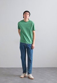 Πράσινη βαμβακερή μπλούζα με κοντά μανίκια και στρογγυλή λαιμόκοψη, συνδυασμένη με μπλε τζιν παντελόνι και μπεζ αθλητικά παπούτσια. Απαλή υφή, άνετη εφαρμογή.