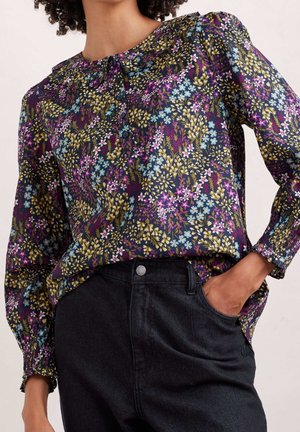 Floral blouse in dark fabric features purple, blue, yellow, and white flowers. Long sleeves with elastic cuffs. Paired with dark jeans.