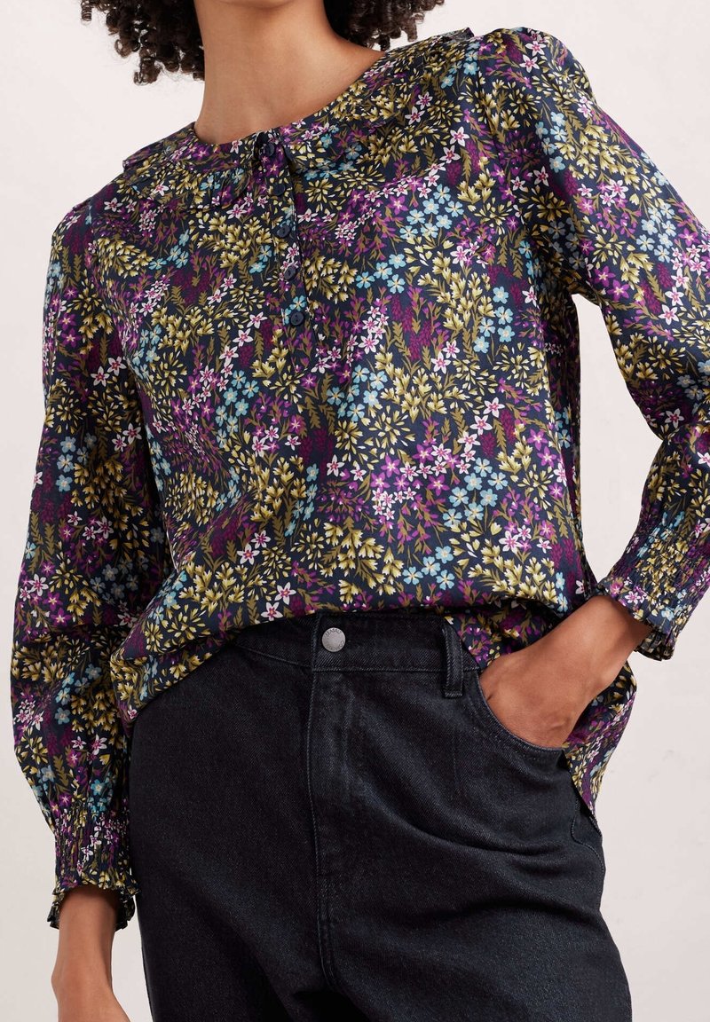 Blomstret bluse i mørkt stof med lilla, blå, gule og hvide blomster. Lange ærmer med elastiske manchetter. Sættes sammen med mørke jeans.
