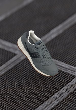 Szary but sportowy Saucony z białą podeszwą i gumową podeszwą zewnętrzną, sznurowany i umieszczony na tle metalowej siatki podłogowej.