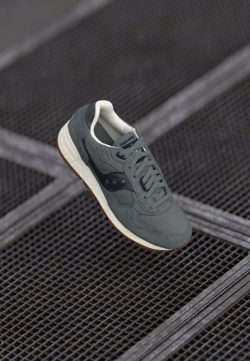 Baskets Saucony grises avec semelle blanche et semelle extérieure en caoutchouc, lacées et positionnées au-dessus d’un sol en grille métallique.