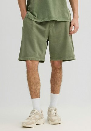 Hombre con pantalones cortos verde oliva hasta la rodilla, con las manos en los bolsillos, calcetines blancos hasta el tobillo y zapatillas beige sobre un fondo claro y liso.