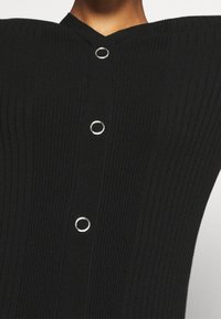 Haut en tricot côtelé noir avec encolure en V et boutons circulaires argentés le long du devant. La texture est lisse et ajustée.