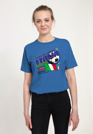 Młoda kobieta ubrana w niebieską koszulkę piłkarską Włoch z nadrukiem piłki, włoskiej flagi oraz lat mistrzostw z przodu.